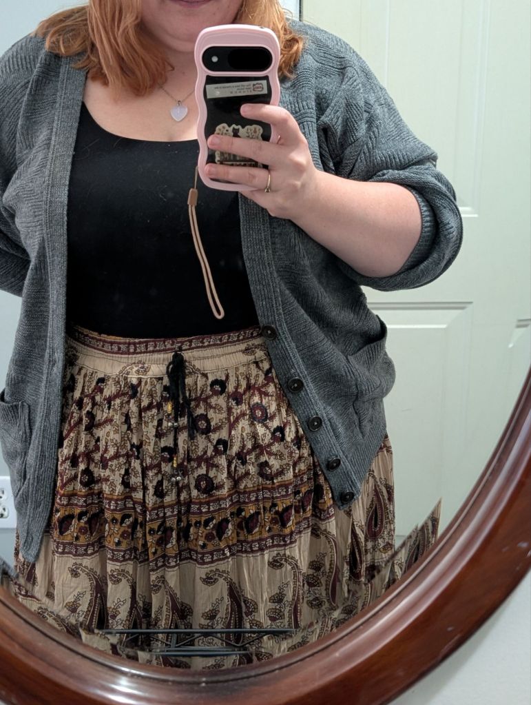 Girl wearing a grey cardigan, a black top and a skirt with a varity of brown, red, yellow and dark blue. 